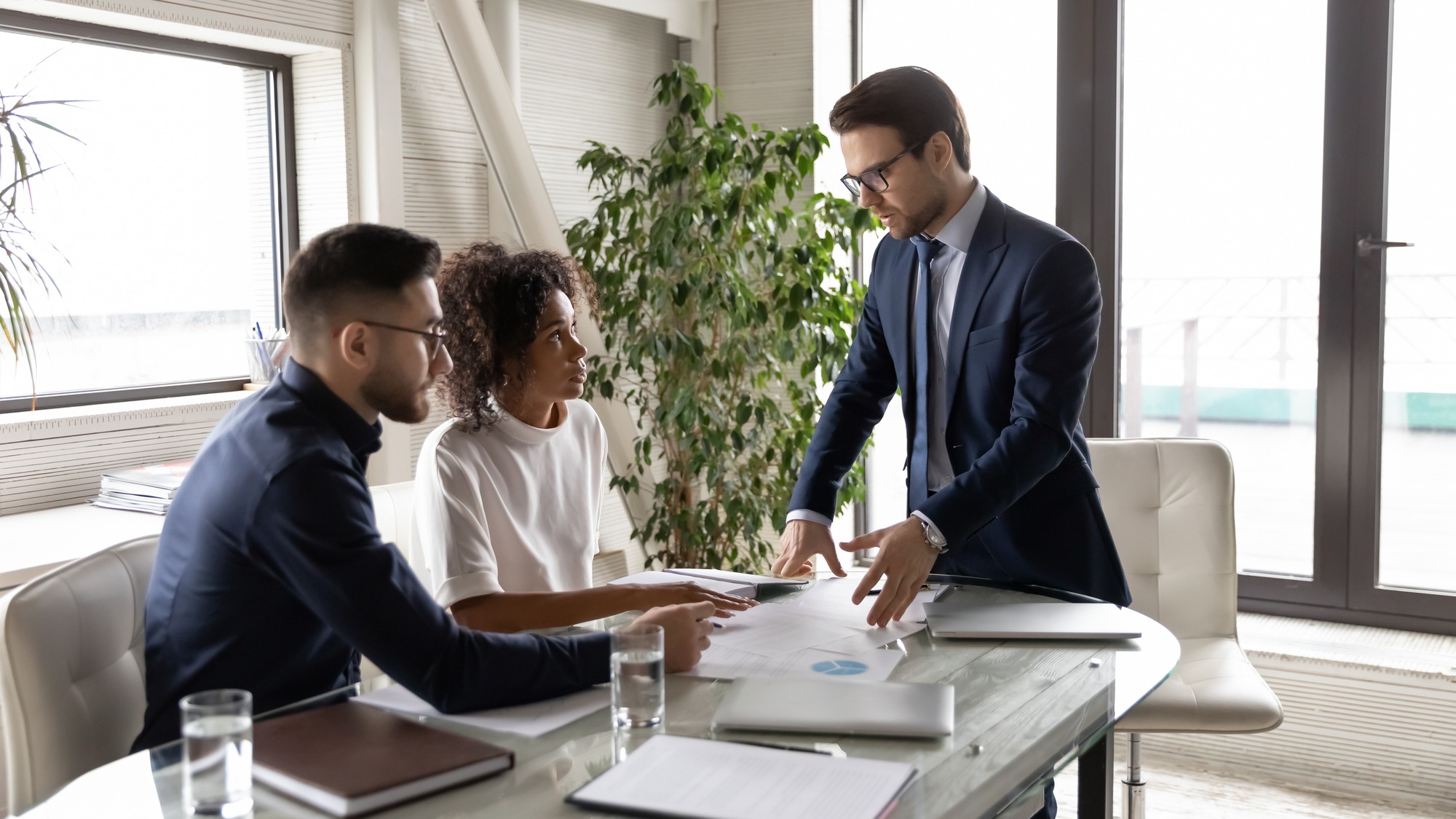 Concentrated multiracial businesspeople gather at desk in boardroom brainstorm over financial paperwork, diverse international colleagues talk discuss business project at meeting in office together.